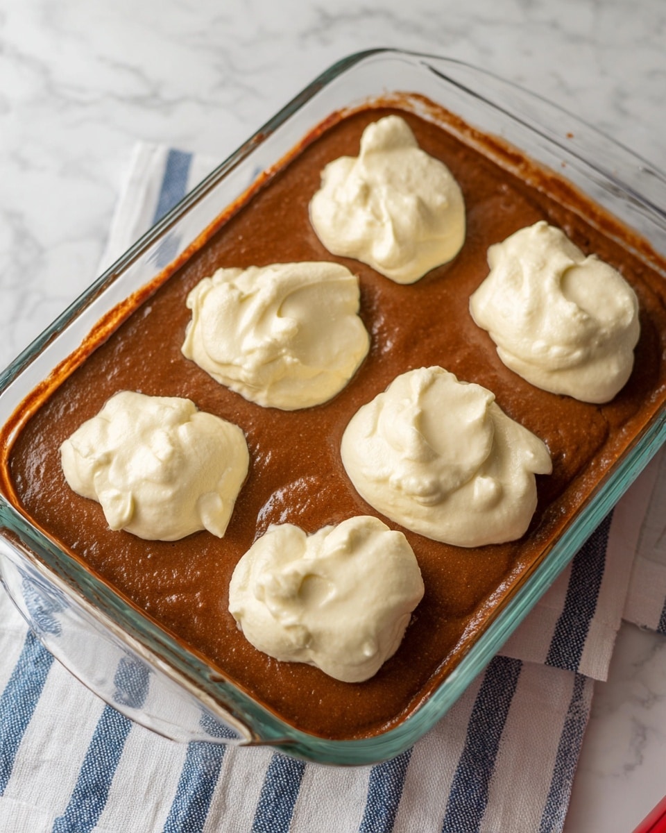 The image shows a clear rectangular glass baking dish filled with a smooth, rich brown batter evenly spread at the bottom as the first layer. On top of this brown batter, there are six dollops of thick, creamy white batter placed in two rows creating a second layer, each dollop slightly rounded and uneven in shape, spaced evenly across the dish. The dish is sitting on a white marbled surface with a striped cloth partially visible underneath the dish. Photo taken with an iphone --ar 4:5 --v 7