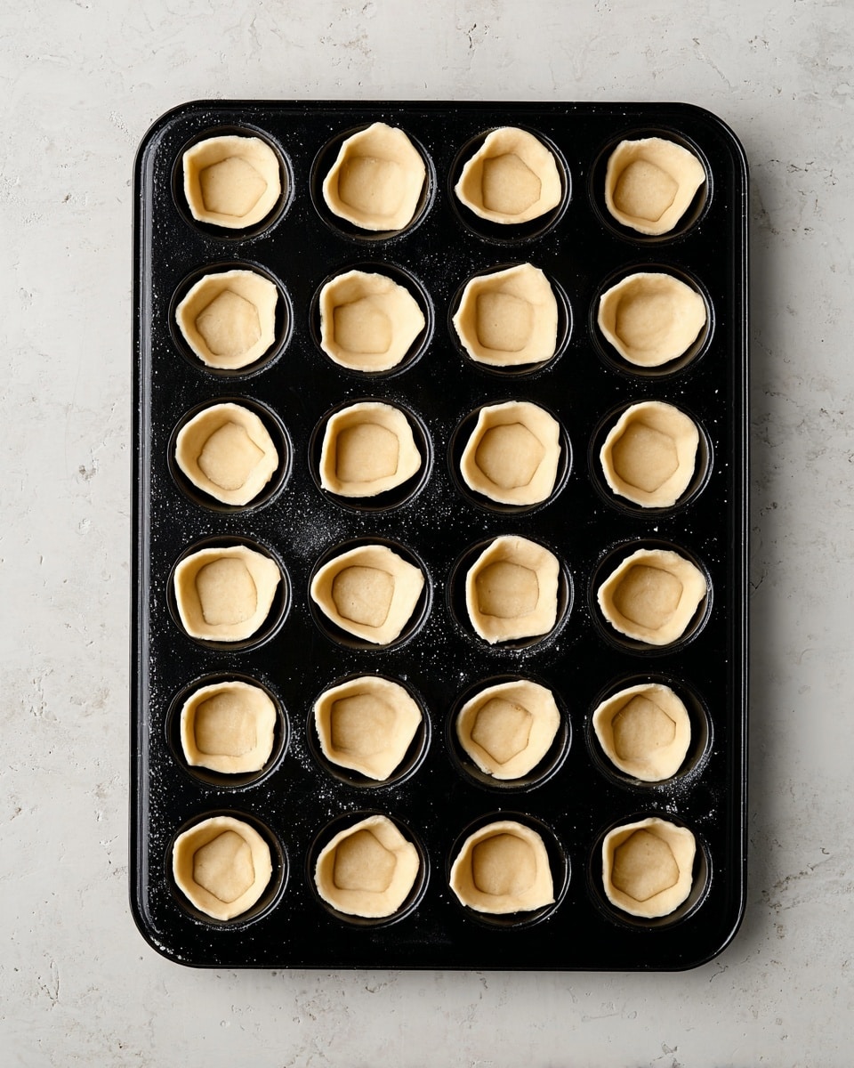 A black mini muffin baking tray with 24 small, raw dough cups inside each section. Each dough cup is pressed down in the center with slightly raised edges, showing a pale beige color and soft texture. The tray sits on a white marbled surface, and there is no other food or decoration visible. photo taken with an iphone --ar 4:5 --v 7