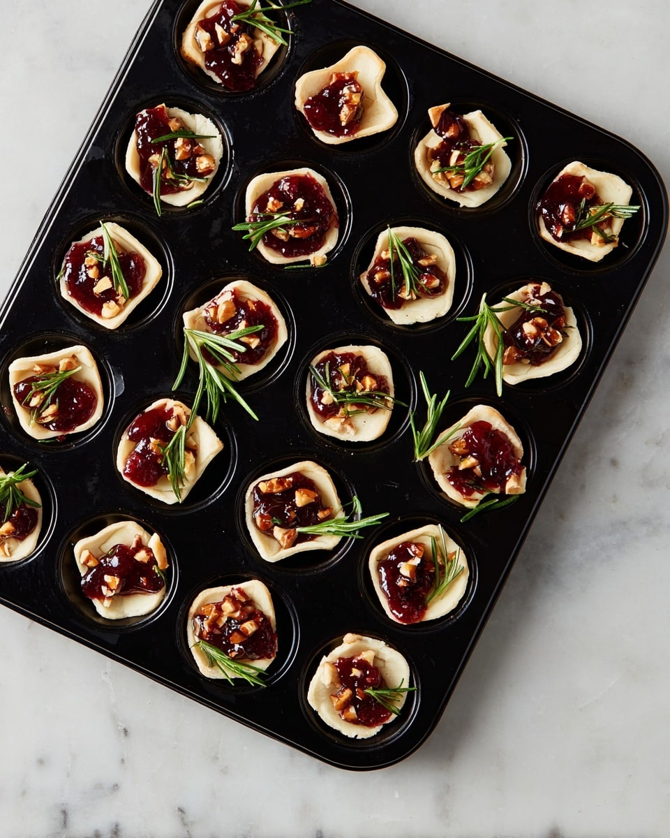 A black muffin tray holds 24 small tartlets with three layers each. The bottom layer is light beige dough shaped into small cups. On top of the dough is a creamy off-white layer, and this is topped with a dark red sauce or jelly. Small brown nut pieces are scattered over the red layer, and green sprigs of rosemary lay across each tartlet. The tray is placed on a white marbled surface. photo taken with an iphone --ar 4:5 --v 7