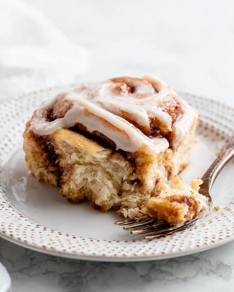 A single slice of cinnamon roll with visible layers of soft, light brown dough swirled with darker cinnamon filling, topped with a creamy white icing that drips slightly over the edges. The texture looks fluffy and slightly dense, with a glossy finish from the icing. The slice sits on a white plate with a dotted pattern around the rim, placed on a white marbled surface. A fork with some torn pieces of the cinnamon roll rests beside it, showing the soft texture inside. Photo taken with an iphone --ar 4:5 --v 7