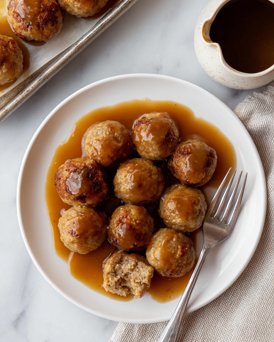The image shows a white plate with nine small, round meatballs arranged in a roughly circular pattern. Each meatball is light brown with a slightly rough texture, and all are covered in a shiny brown gravy that pools around the base of the meatballs on the plate. One meatball near the edge is broken open, revealing a lighter inside, and a silver fork is resting on the plate next to it. The plate sits on a white marbled surface, with part of a baking tray holding more meatballs visible in the upper left corner and a small ceramic jug filled with dark brown gravy resting on the surface in the upper right corner. photo taken with an iphone --ar 4:5 --v 7