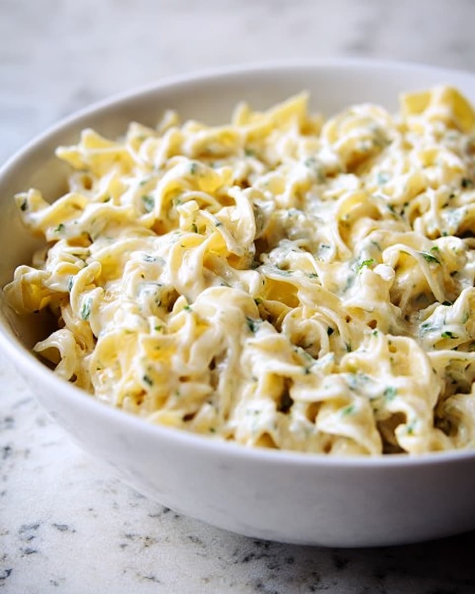 The image shows a close-up of a white bowl filled with creamy pasta. The pasta is twisted and ribbon-like, coated in a thick, white sauce with small green bits, likely herbs, scattered evenly throughout. The texture of the sauce looks smooth and rich, covering each curl of the pasta well. The bowl is set on a white marbled surface, and the overall look is simple and fresh without extra toppings or garnishes. Photo taken with an iphone --ar 4:5 --v 7