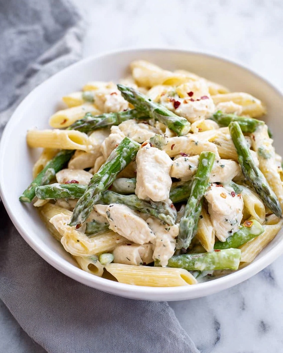 The image shows a white bowl filled with a creamy pasta dish. The dish has three main layers: the base layer is light yellow penne pasta, followed by chunks of white cooked chicken pieces scattered throughout. On top of these, bright green asparagus tips are evenly spread. The pasta and chicken are coated with a smooth, white creamy sauce, and small flecks of black pepper and red pepper flakes add subtle color accents. The bowl rests on a white marbled surface, with a soft grey cloth slightly visible behind it. photo taken with an iphone --ar 4:5 --v 7