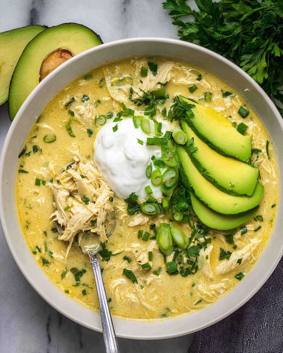 A white bowl is filled with a creamy yellow soup containing shredded pieces of chicken and small bits of green herbs mixed evenly throughout. On top, there is a dollop of white sour cream in the center. Around the sour cream, thin slices of fresh avocado with light green and yellow tones are arranged in a fan shape on one side. Chopped green onions and finely chopped cilantro are sprinkled over the soup, avocado, and sour cream, adding a bright green contrast. A spoon is dipped into the soup on the left side of the bowl, scooping up some of the creamy chicken mixture. The bowl is set on a white marbled surface with two slices of avocado and some parsley scattered nearby. photo taken with an iphone --ar 4:5 --v 7