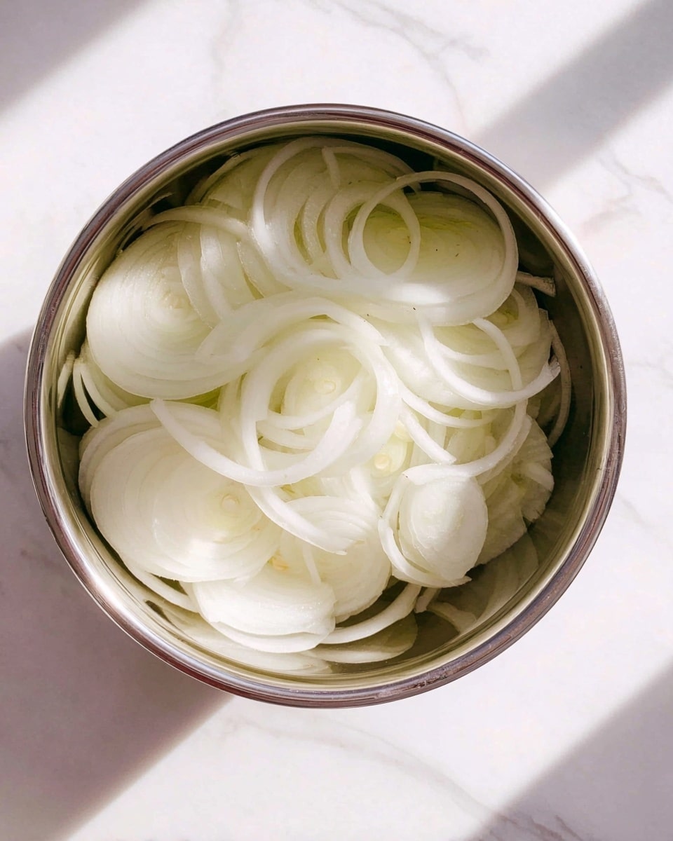 A shiny silver metal bowl filled with several layers of thinly sliced white onions, showing rings and crescent shapes overlapping each other. The bowl is placed on a white marbled surface, and the onions inside have a smooth, slightly translucent texture with light cream and pale yellow shades. Photo taken with an iphone --ar 4:5 --v 7