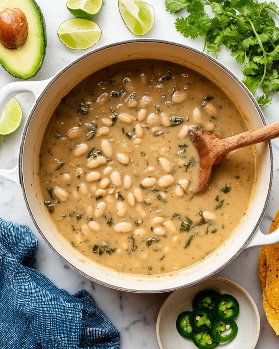 A large white pot filled with creamy beige soup dotted with soft white beans and small bits of greens, with a wooden spoon resting inside the pot. Around the pot on a white marbled surface are fresh lime wedges, bright green cilantro leaves, a halved avocado showing a seed, sliced green jalapeno on a small white plate, a folded blue cloth napkin, and a piece of crispy tortilla. Photo taken with an iphone --ar 4:5 --v 7