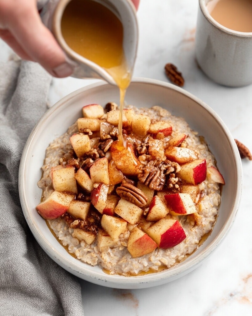 A close-up view of a bowl of oatmeal topped with small chunks of cooked apples and sprinkled with chopped pecans. The oatmeal forms the bottom layer with a creamy, soft texture in a neutral beige color, filling the bowl. On top sits a generous layer of warm apple pieces with red skin and light brown cinnamon dusting, creating a mix of reds and browns with a slightly glossy look. Scattered across the apples are small pieces of pecans in light and dark brown tones with a rough texture. From above, a woman's hand pours a stream of golden syrup over the apples and nuts. The bowl is white and sits on a white marbled surface, with a soft grey cloth and a cup of light brown liquid visible near the edges. Photo taken with an iphone --ar 4:5 --v 7