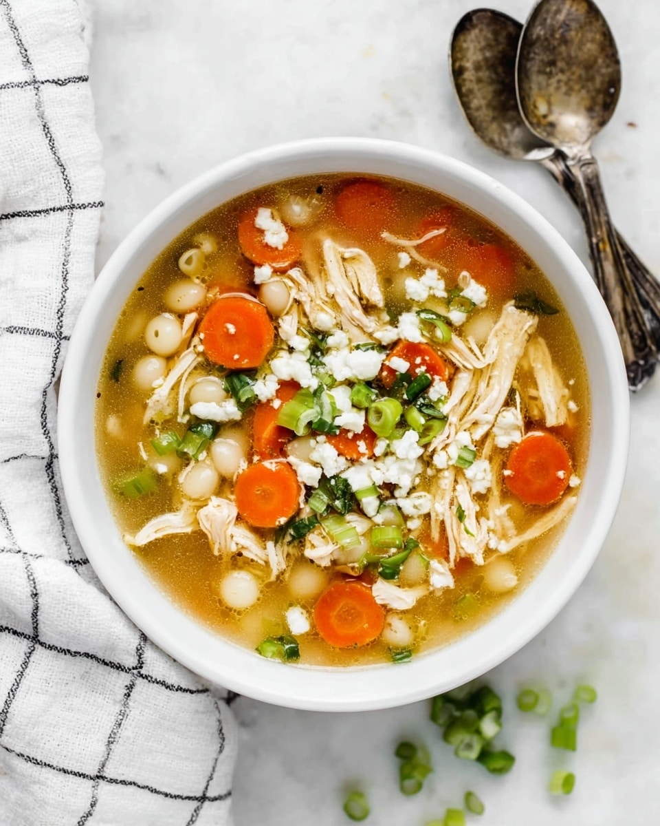 A white bowl filled with clear broth soup showing visible layers of shredded light beige chicken at the bottom, round bright orange carrot slices scattered throughout, small white pearl-like pasta spread evenly, and topped with white crumbled cheese and bright green chopped scallions. The bowl is placed on a white marbled surface with some green scallion pieces scattered around, a white cloth with black grid lines partially underneath on the left side, and two rustic silver spoons resting on the right. photo taken with an iphone --ar 4:5 --v 7