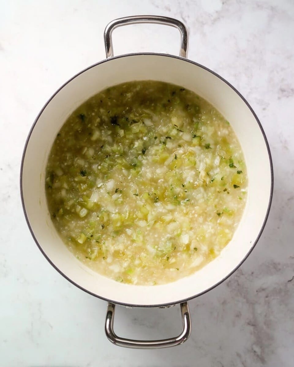 The image shows a white cooking pot with two metal handles on a white marbled surface. Inside the pot, there is a single layer of steaming, soft mixture of light green and yellow ingredients, with small bits of onions and herbs visible. The texture looks thick and slightly chunky, spreading evenly across the pot's bottom. Photo taken with an iphone --ar 4:5 --v 7