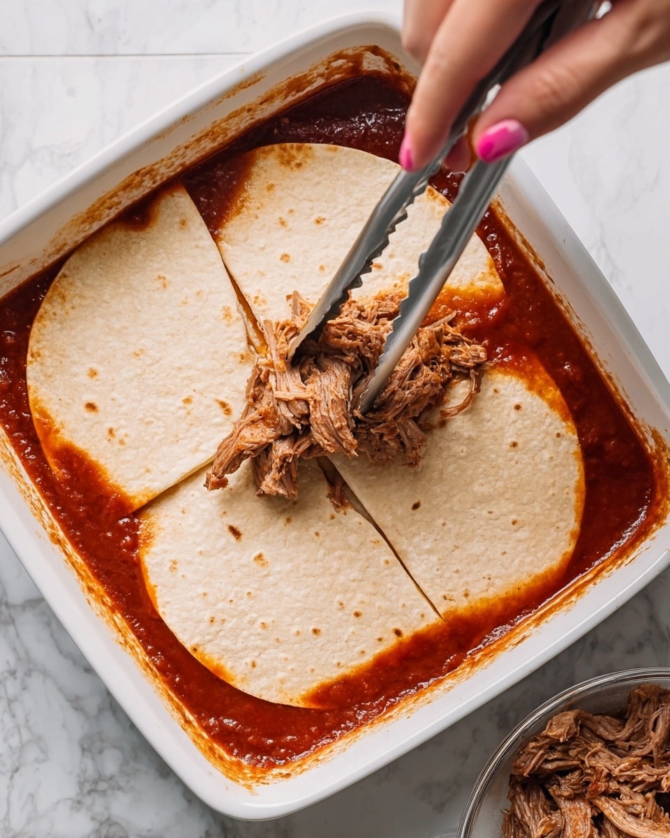 The photo shows a white square baking dish with a shallow layer of red sauce at the bottom. On top of the sauce, there are four broken pieces of light beige tortillas that cover most of the sauce. A woman's hand with pink nail polish is holding metal tongs, lifting a chunk of shredded brown meat above the tortillas. In the bottom right corner, there is a blurry look of a bowl with brown shredded meat, indicating more filling ready to be placed. The whole scene is set on a white marbled surface. photo taken with an iphone --ar 4:5 --v 7