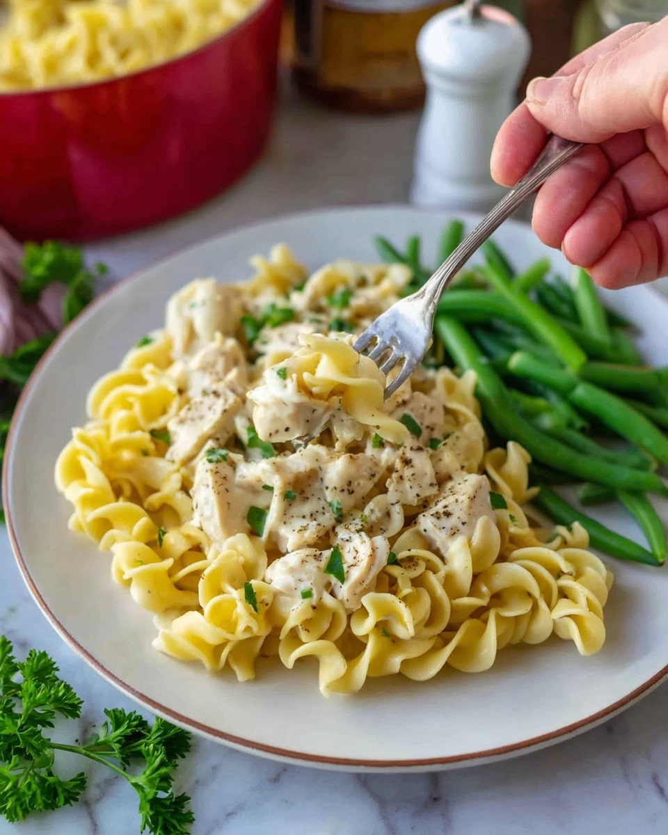 A white plate holds a serving of three layers: the bottom layer is light yellow twisted egg noodles with a soft, slightly creamy texture; the middle layer has small pieces of light beige chicken mixed into the noodles, sprinkled with black pepper and green herbs; the top layer features bright green cooked green beans on the right side, with a woman's hand holding a fork lifting a bite of noodles and chicken from the left side. The plate sits on a white marbled surface with some fresh green parsley leaves near the bottom, and in the blurred background, there are white salt and pepper shakers and a red bowl filled with more noodles. Photo taken with an iphone --ar 4:5 --v 7