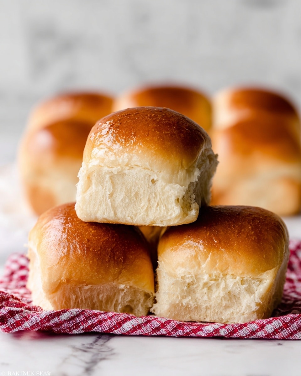Five soft, golden brown bread rolls are stacked on a red and white checkered cloth on a white marbled surface. The textured tops of the rolls are shiny and smooth, while the torn edges reveal a fluffy, light-colored inside. The rolls are arranged with three on the bottom and two on top, showing their airy and tender crumb. In the blurred background, more bread rolls can be seen, all with a warm and fresh appearance. photo taken with an iphone --ar 4:5 --v 7