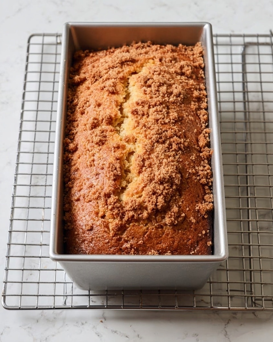 A golden brown loaf cake sits in a simple silver metal rectangular pan with straight sides. The cake's textured top is uneven with small cracks and a sprinkled light brown crumb topping adding a rough, crunchy look. The pan rests on a metal cooling rack with a grid pattern, placed on a white marbled surface that has soft gray veins. The overall scene looks bright and clean, focusing on the warm tones of the freshly baked cake photo taken with an iphone --ar 4:5 --v 7