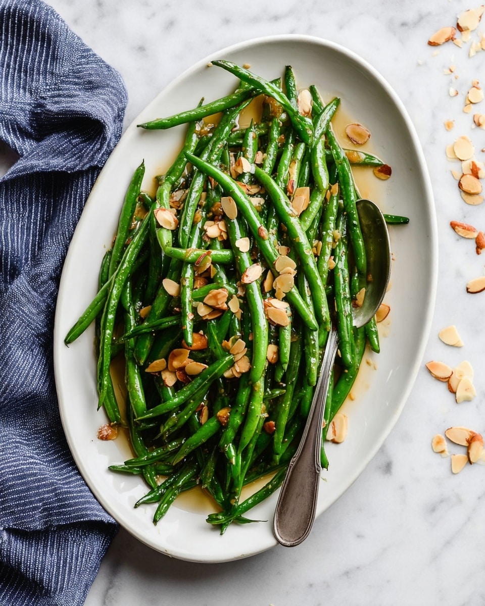 A large white oval plate filled with shiny green beans that are thin and long, topped with toasted almond slices scattered evenly over them. The beans look lightly cooked and glistening with oil or butter. A silver spoon rests on the right side of the plate among the beans. Around the plate, on the white marbled surface, a few almond slices are spread loosely. A woman’s hand wearing a blue and white striped cloth is positioned on the left side next to the plate. photo taken with an iphone --ar 4:5 --v 7
