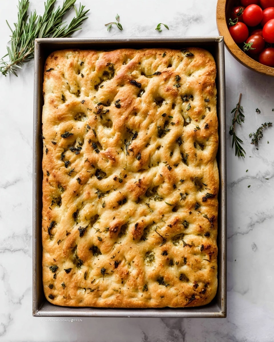 A rectangular focaccia bread with a bumpy, golden-brown crust sits inside a metal baking pan, its surface dotted with small pockets and churrs. The bread is speckled with bits of dark green herbs and light brown seasoning. Small fresh herb leaves are scattered lightly on top, adding a touch of brightness. The edges are puffed up and slightly more browned than the rest of the flat bread. The pan rests on a white marbled surface with a wooden bowl of red tomatoes and sprigs of rosemary nearby. photo taken with an iphone --ar 4:5 --v 7