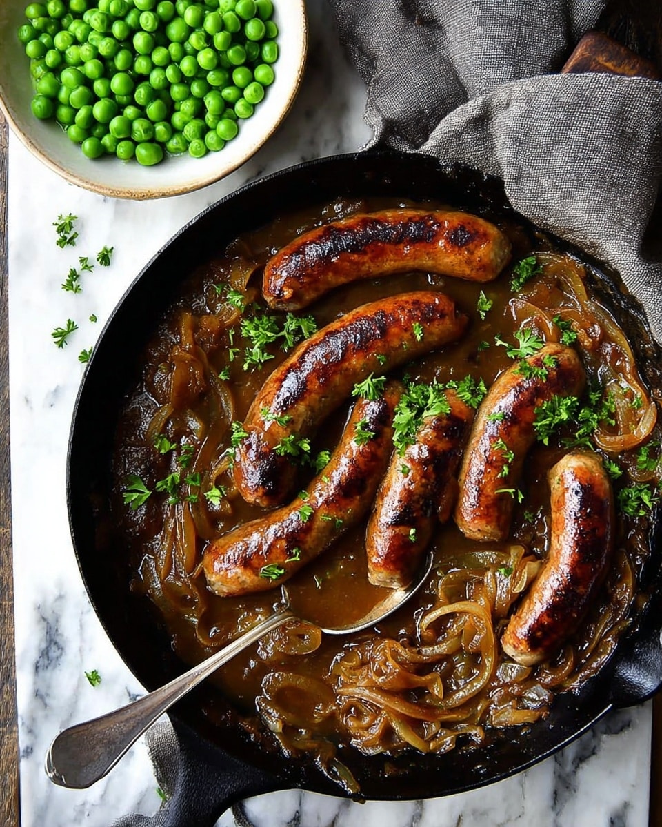 The image shows a black cast iron pan with six browned sausages on top of a rich brown gravy mixed with caramelized onion strands, sprinkled with fresh green parsley. A metal spoon is partly resting on the pan’s edge, dipped into the gravy. Next to the pan is a white bowl filled with bright green peas, all set on a white marbled surface with a folded grey cloth nearby. Photo taken with an iphone --ar 4:5 --v 7