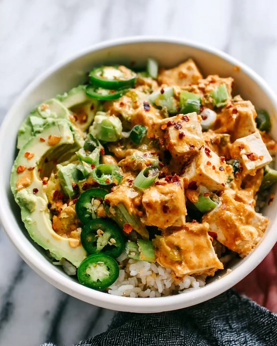 The image shows a white bowl filled with a layered dish on a white marbled surface. At the bottom, there is a layer of white rice. On top of the rice, there are chunks of light brown tofu mixed with bright green avocado pieces and some sliced green jalapeños all around. The dish is covered with a creamy orange sauce with red chili flakes sprinkled over it, adding texture. A woman’s hand is holding the bowl on the right side of the image. The photo is taken with an iphone --ar 4:5 --v 7