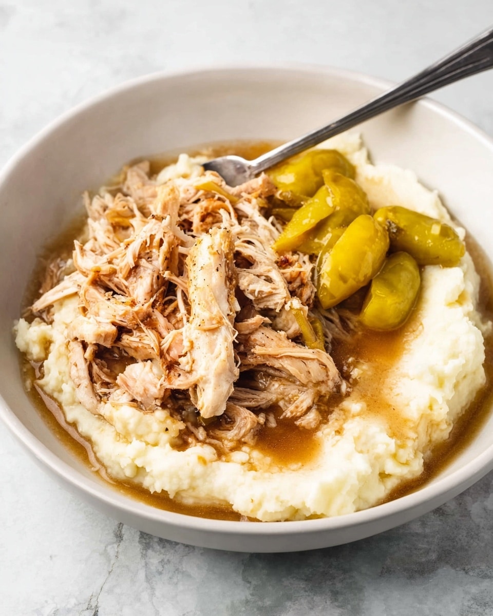 A white bowl holds two main layers: at the bottom, a smooth, creamy layer of white mashed potatoes, topped with a generous portion of shredded light brown chicken mixed with brown gravy. On one side, two whole yellow pepperoncini peppers rest partially on the chicken and mashed potatoes. A silver fork is placed inside the bowl on the left side, resting on the mashed potatoes. The bowl sits on a white marbled textured surface. photo taken with an iphone --ar 4:5 --v 7