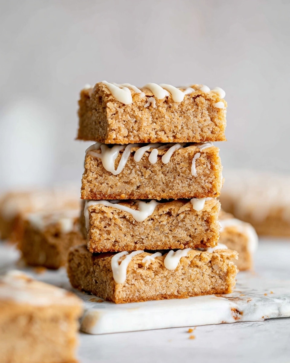 The image shows a stack of four thick, square blondie bars placed on a white marbled surface with some more blondies in the background. Each blondie has a golden brown color with a slightly crumbly and moist texture, and the tops are drizzled with thin lines of white icing. The bars have a dense, soft inside and a lightly crisp edge. The background is blurred and neutral, focusing attention on the stack of blondies. photo taken with an iphone --ar 4:5 --v 7