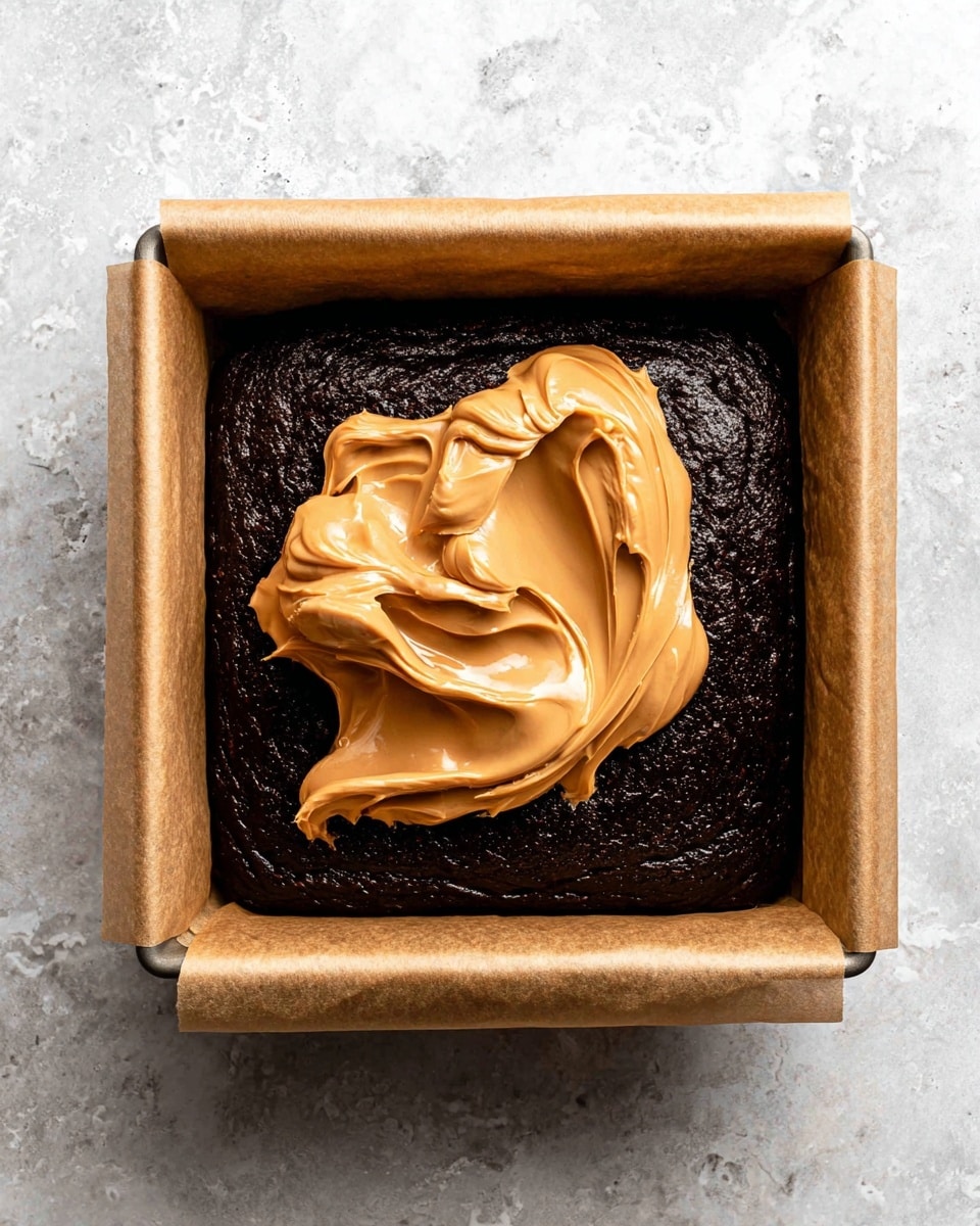 A square baking pan lined with brown parchment paper holds a dark, almost black, rich cake layer that fills the pan completely. On top, there is a large dollop of smooth, creamy peanut butter spread, with swirled peaks and a light brown color sitting in the middle of the cake layer. The pan rests on a white marbled surface. photo taken with an iphone --ar 4:5 --v 7