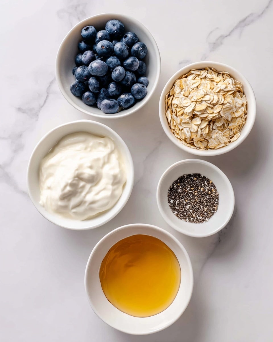 Five small white bowls are placed on a white marbled surface, each holding a different ingredient arranged in a loose cluster. The top left bowl contains dark blue, round blueberries labeled