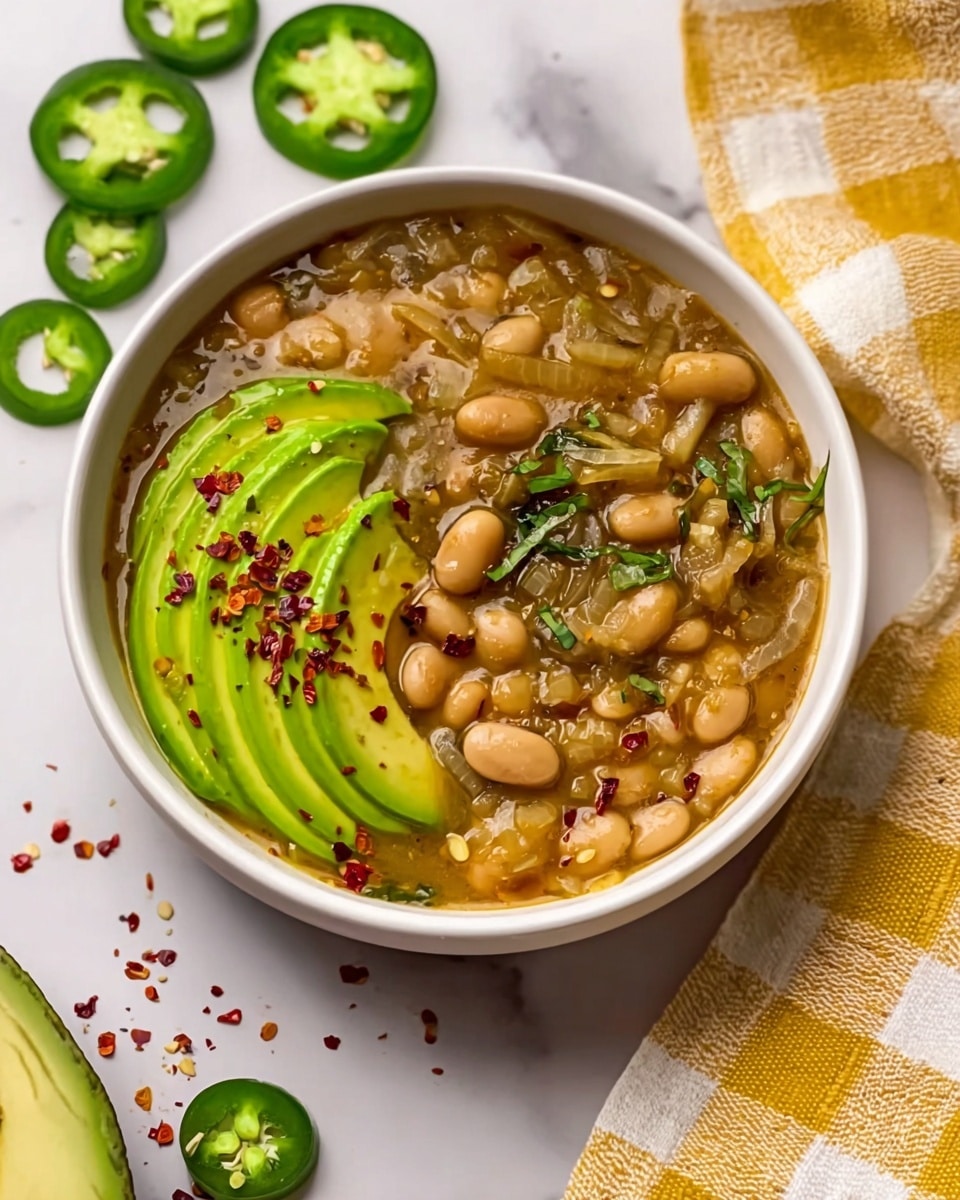 The image shows a white bowl filled with a thick stew made of white beans and sliced jalapeño peppers. The stew has a brownish sauce with visible pieces of cooked onion and small green herbs scattered on top. On one side of the bowl, there are several slices of fresh avocado, bright green and smooth, placed neatly with a sprinkle of red chili flakes adding contrast. Around the bowl, there are sliced jalapeño rings and some red chili flakes scattered on a white marbled surface. A folded yellow and white checkered cloth is placed next to the bowl. Photo taken with an iphone --ar 4:5 --v 7