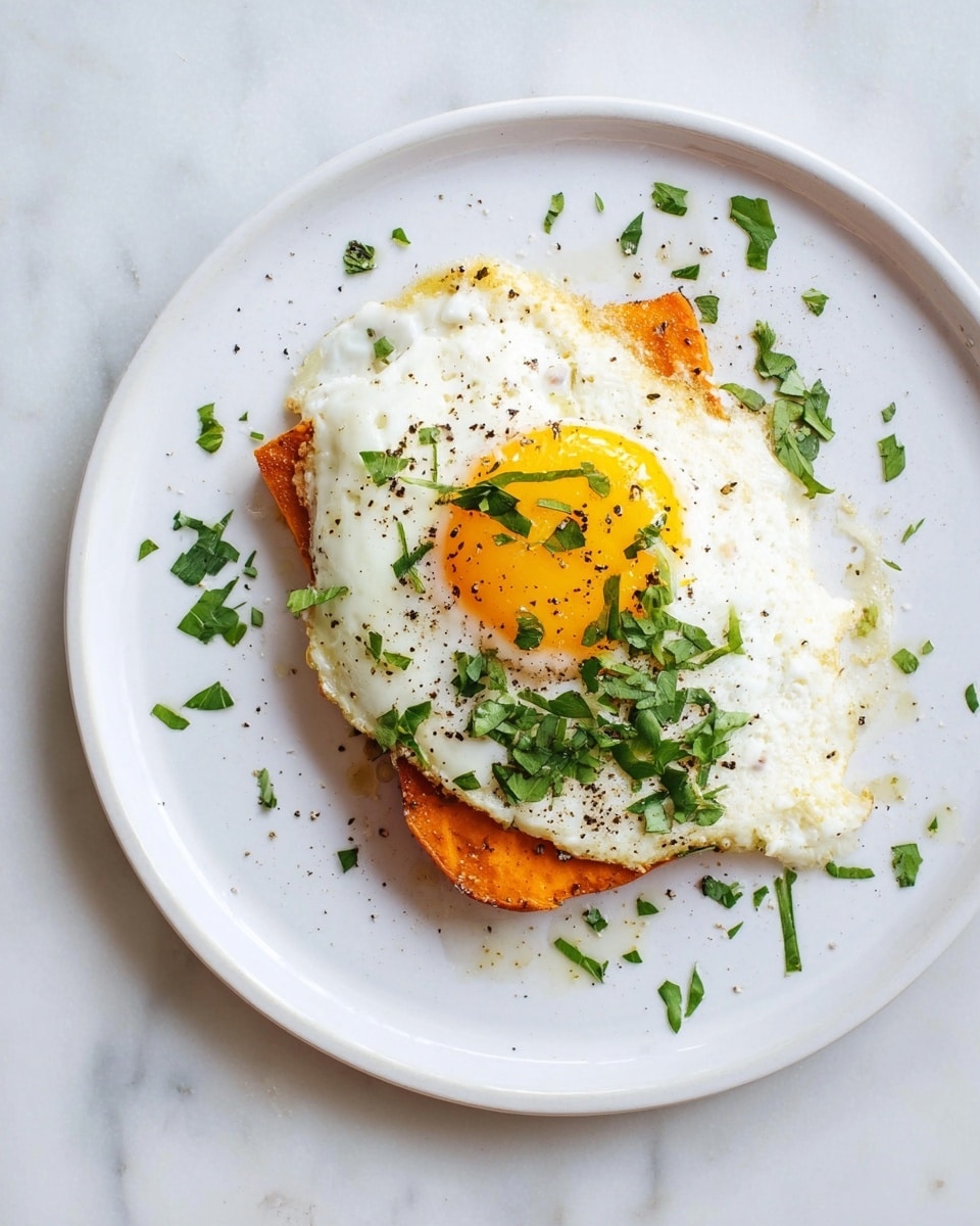 A white plate holds a simple dish with two layers: the bottom layer is a rectangular piece of roasted orange sweet potato with slightly browned edges, and on top sits a fried egg with a bright yellow runny yolk and white cooked edges that spread slightly beyond the sweet potato. The egg is sprinkled with black pepper and fresh chopped green herbs scattered on and around it. The plate is placed on a white marbled surface. photo taken with an iphone --ar 4:5 --v 7