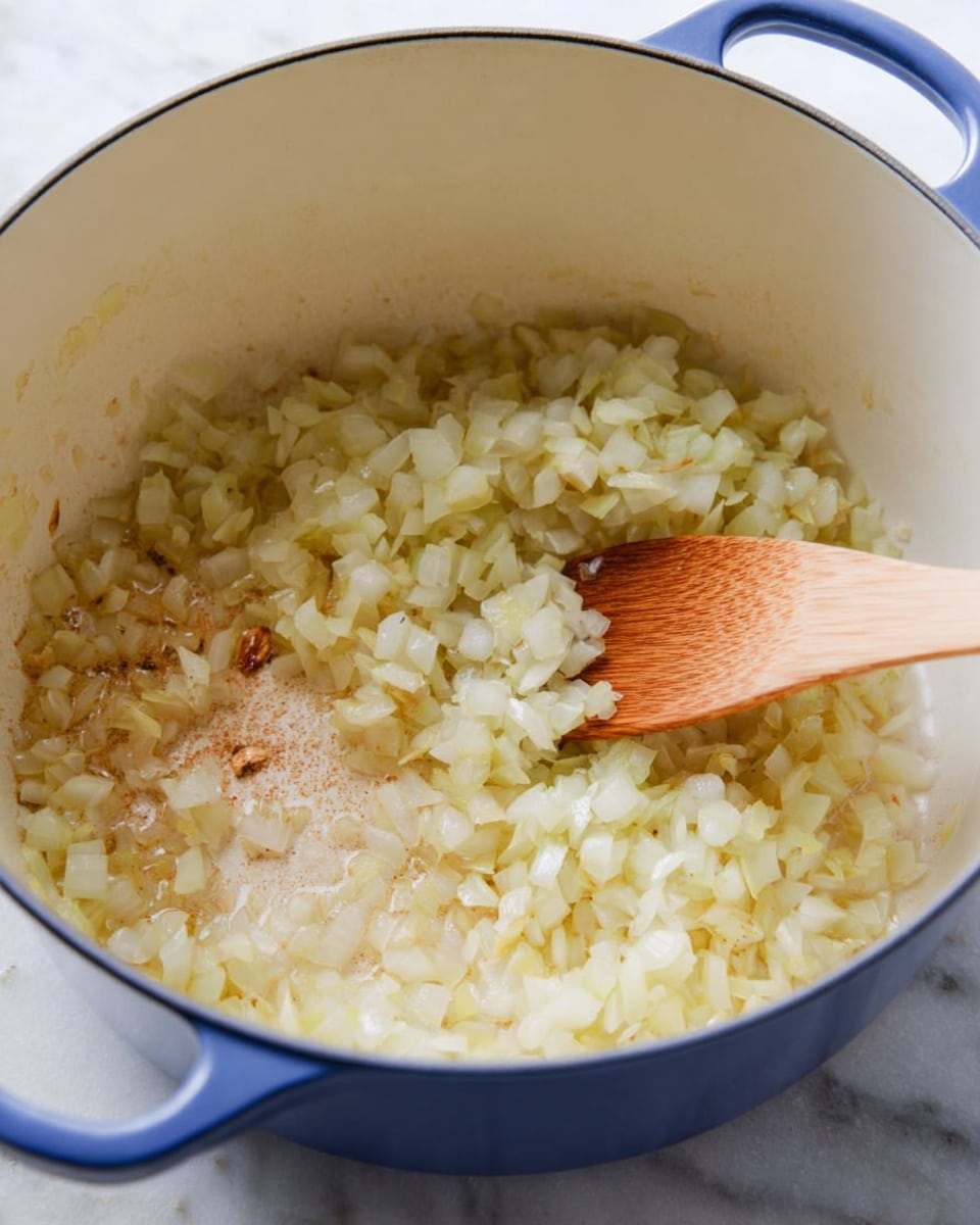 A close-up view inside a white cooking pot with a blue outer edge, showing finely chopped translucent onions being stirred with a light brown wooden spoon. The onions have a soft, slightly cooked look with a pale yellow and white texture. Some light brown bits are scattered on the bottom of the pot, indicating light cooking. The pot is placed on a white marbled surface, and the overall scene has soft, natural lighting. Photo taken with an iphone --ar 4:5 --v 7