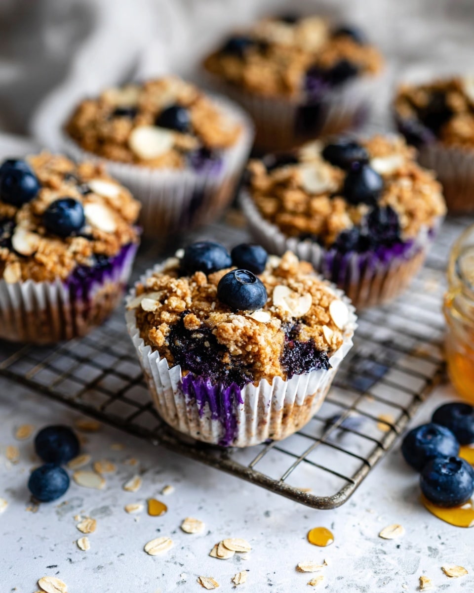 The image shows several blueberry oatmeal muffins, each in white paper liners with a purple tint visible underneath. The muffins have a golden brown oatmeal base dotted with plump, dark blueberries throughout. On top, there is a rough oat crumb layer with scattered sliced almonds and whole blueberries adding texture and color contrast. The muffins rest on a metal cooling rack placed over a white marbled texture, with a few loose blueberries and drops of honey scattered around. The background is softly blurred, emphasizing the warm, crumbly texture and deep blue highlights of the muffins. Photo taken with an iphone --ar 4:5 --v 7