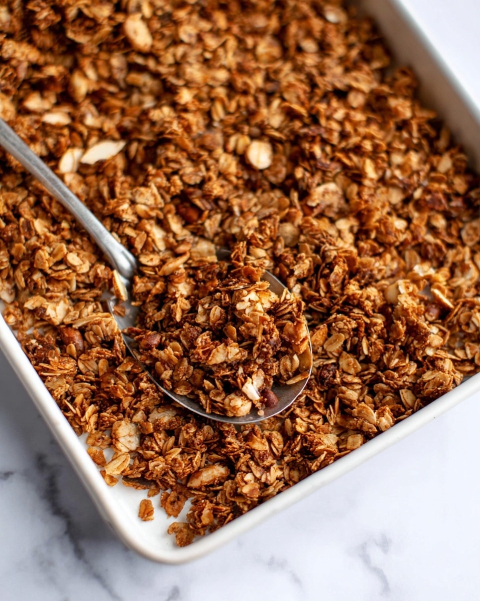 The image shows a close-up of a white baking tray filled with a thick layer of toasted granola. The granola has a mix of small and medium clusters, featuring oats and chopped nuts with a golden to dark brown color, showing a crunchy texture. A silver spoon is scooping some granola from the tray, positioned slightly off-center to the right. The tray rests on a white marbled surface, creating a clean and bright background. photo taken with an iphone --ar 4:5 --v 7