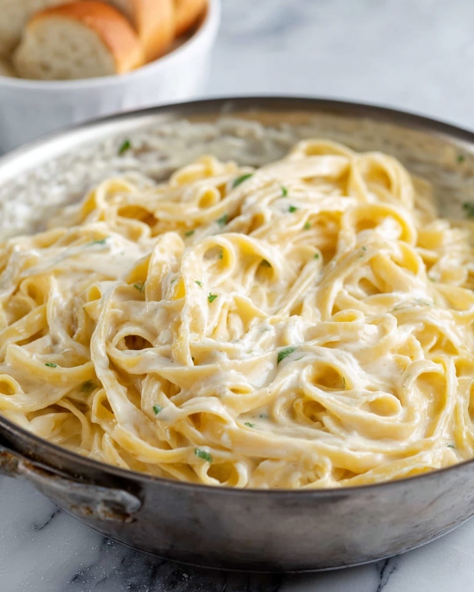 The image shows a pan filled with creamy fettuccine pasta. The pasta noodles are pale yellow and coated evenly in a smooth, creamy white sauce with a few tiny green herb specks scattered throughout. The pasta is twisted and layered loosely inside the pan. The background is a white marbled texture, and part of a white bowl with bread slices is visible in the upper background. photo taken with an iphone --ar 4:5 --v 7