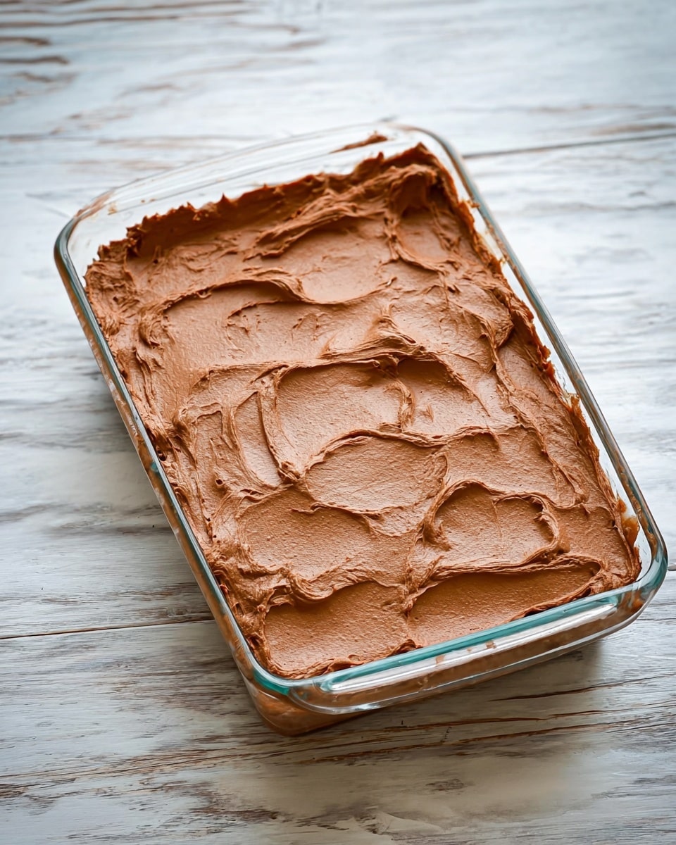 A glass rectangular dish filled with one thick layer of smooth, creamy, light brown chocolate frosting spread evenly but with visible swirls and peaks on the top. The dish is placed on a white marbled surface with light wood texture visible in the background. The frosting looks soft and rich, covering the entire surface of the dish fully, with some textured strokes giving it a whipped look. photo taken with an iphone --ar 4:5 --v 7
