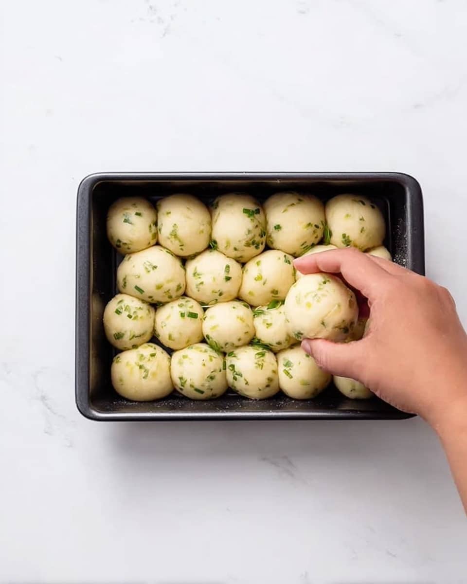 A rectangular black baking pan is filled with small, round dough balls placed closely together in two layers. The dough balls are pale yellow with green herb pieces sprinkled evenly on their smooth surface. A woman's hand is gently placing another dough ball on top, near the right side of the pan. The pan sits on a white marbled surface, creating a clean and bright background. photo taken with an iphone --ar 4:5 --v 7