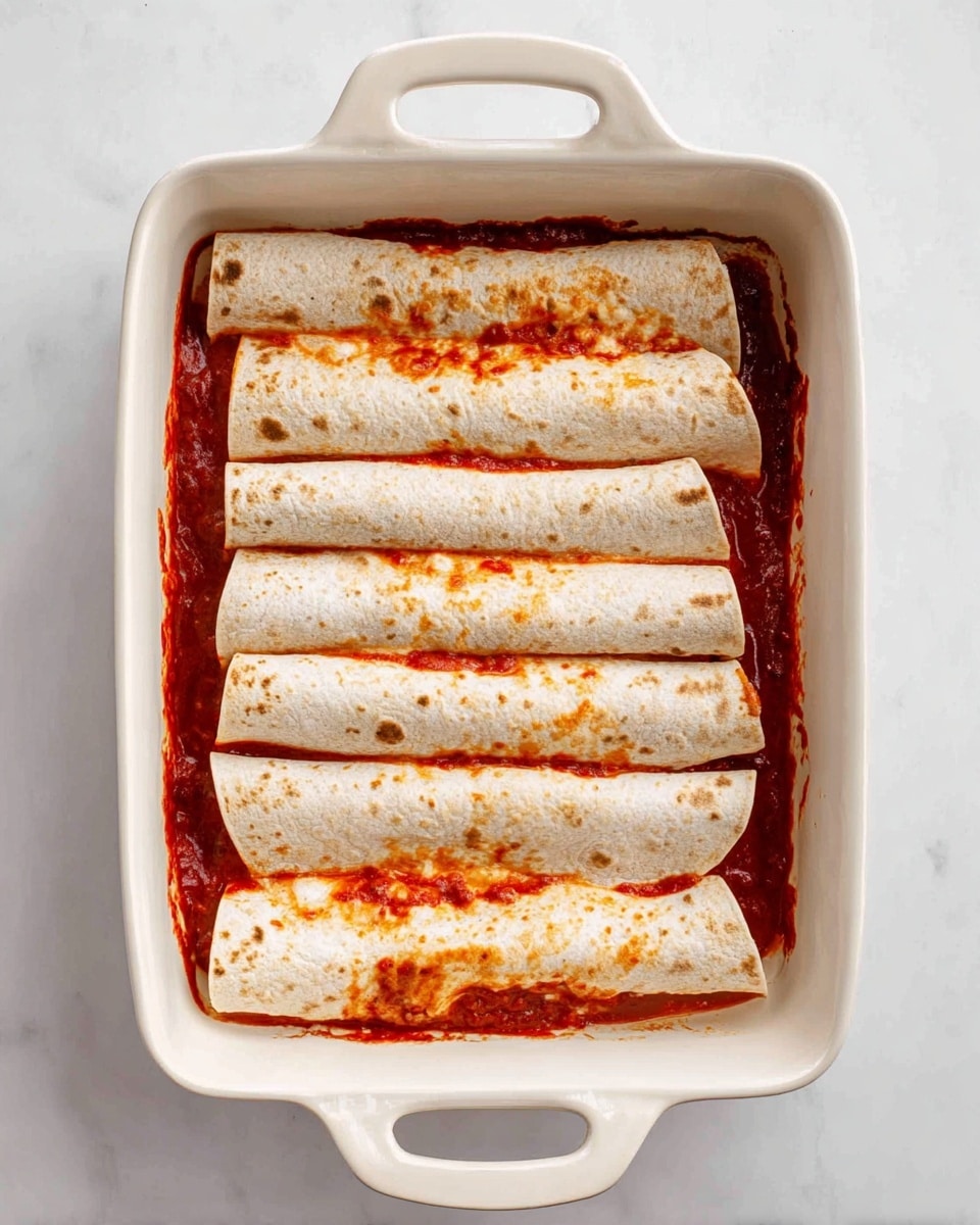 The image shows a white rectangular baking dish with handles on both ends, filled with six rolled tortillas laid side by side. The tortillas are pale with a few light brown spots and are covered in a red sauce, which can be seen in the gaps around and between the tortillas and slightly on top. The dish is placed on a white marbled textured surface. Photo taken with an iphone --ar 4:5 --v 7