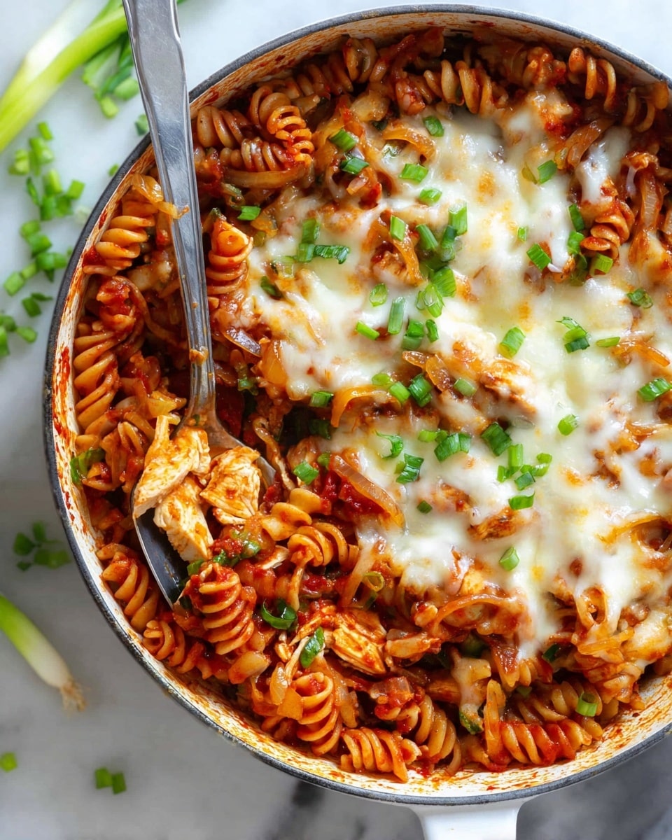 The image shows a white pan filled with cooked spiral pasta covered in red tomato sauce, mixed with pieces of cooked chicken and sautéed onions. On top of the pasta, there is a layer of melted white cheese with some browned spots where it melted in the oven. Fresh green chopped spring onions are sprinkled over the cheese. A silver spoon is inside the pan showing some pasta and chicken, and the whole pan is placed on a white marbled surface with a few green onion pieces scattered around. photo taken with an iphone --ar 4:5 --v 7