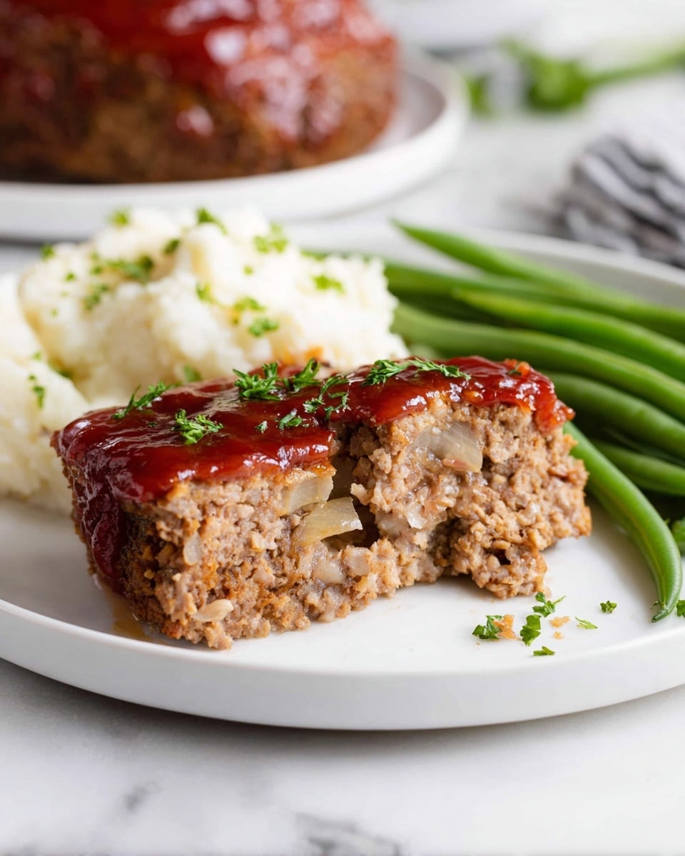 A white plate holds a meal with three main layers: in the front are two slices of meatloaf, one partially eaten to show a soft, slightly crumbly texture mixed with small pieces of onion inside. The top of the meatloaf is covered with a thick, shiny red glaze and small green parsley leaves scattered on top. Behind the meatloaf, on the right side, bright green steamed green beans are stacked neatly. On the left side of the plate, there is a serving of fluffy white mashed potatoes with a soft texture. The plate sits on a white marbled surface, and in the background, another whole meatloaf covered with the same red glaze is slightly out of focus. Photo taken with an iphone --ar 4:5 --v 7