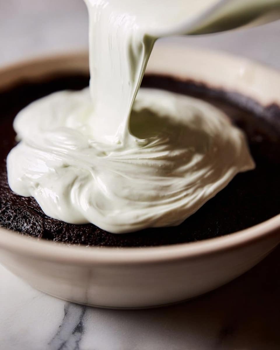 A close-up image shows a white creamy layer being poured thickly over a dark brown base in a white bowl. The creamy layer is smooth and soft with visible folds and swirls as it flows out. The dark brown base looks dense and moist, filling the bottom part of the bowl. The bowl sits on a white marbled textured surface, and the background is softly blurred. photo taken with an iphone --ar 4:5 --v 7