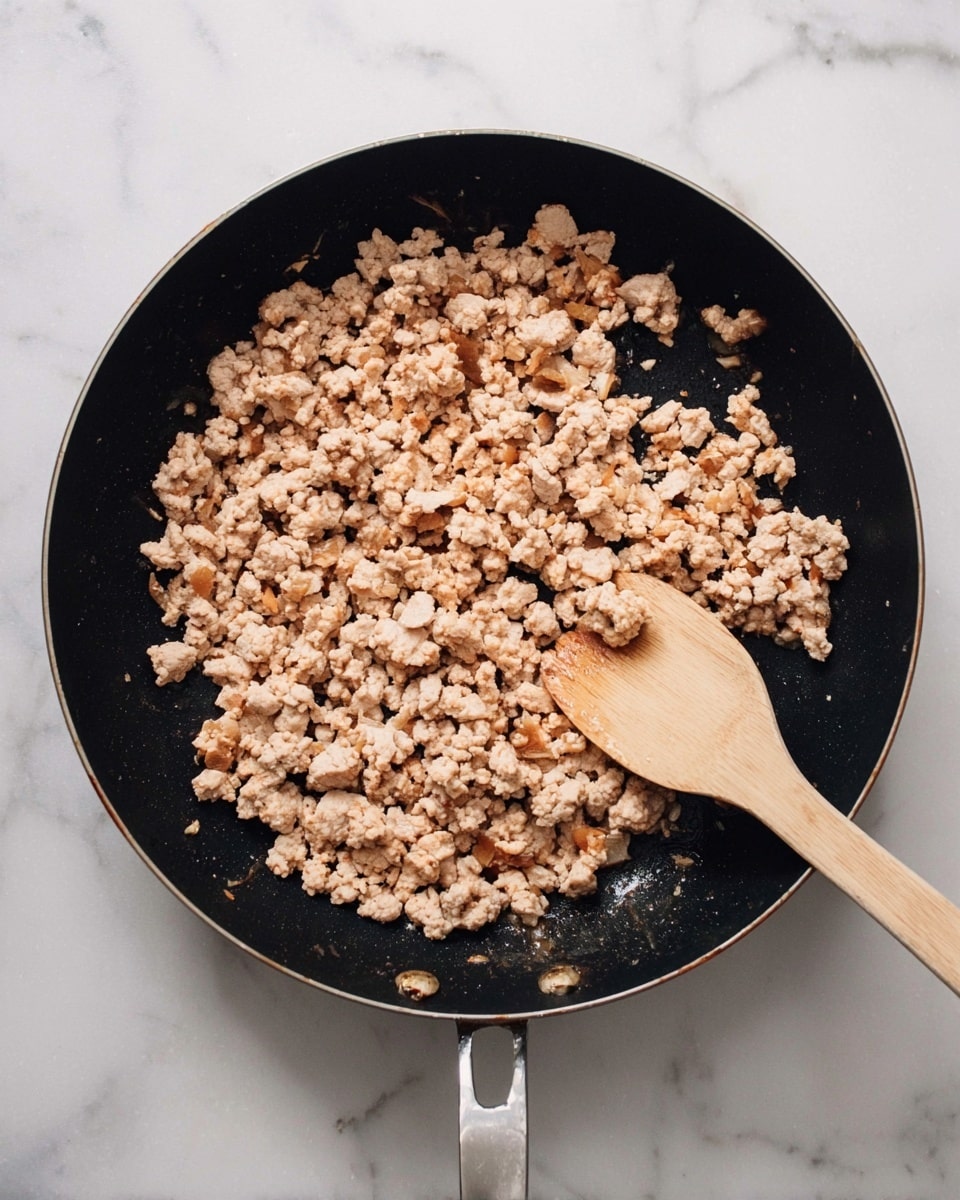 A black frying pan filled with cooked ground meat that is light brown with small bits of slightly darker browned pieces. The meat has a crumbly texture and is spread evenly in the pan. Inside the pan is a wooden spoon with a pale wood color resting on the right side. The pan is placed on a white marbled surface. photo taken with an iphone --ar 4:5 --v 7