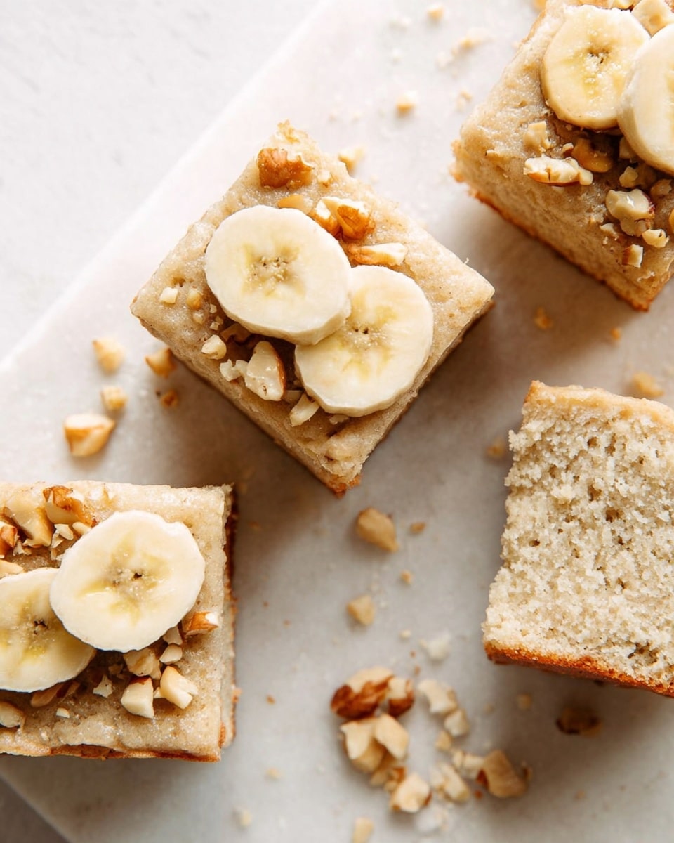 The image shows square pieces of banana cake with a light beige color and soft texture. Each piece has two banana slices on top, placed near the center, and sprinkled with small chopped nuts that add a rough texture and golden-brown color. The cake pieces are arranged on a white marbled surface, with some nut crumbs scattered around. The inside of the cake has a spongy and airy look, with small air bubbles visible. photo taken with an iphone --ar 4:5 --v 7