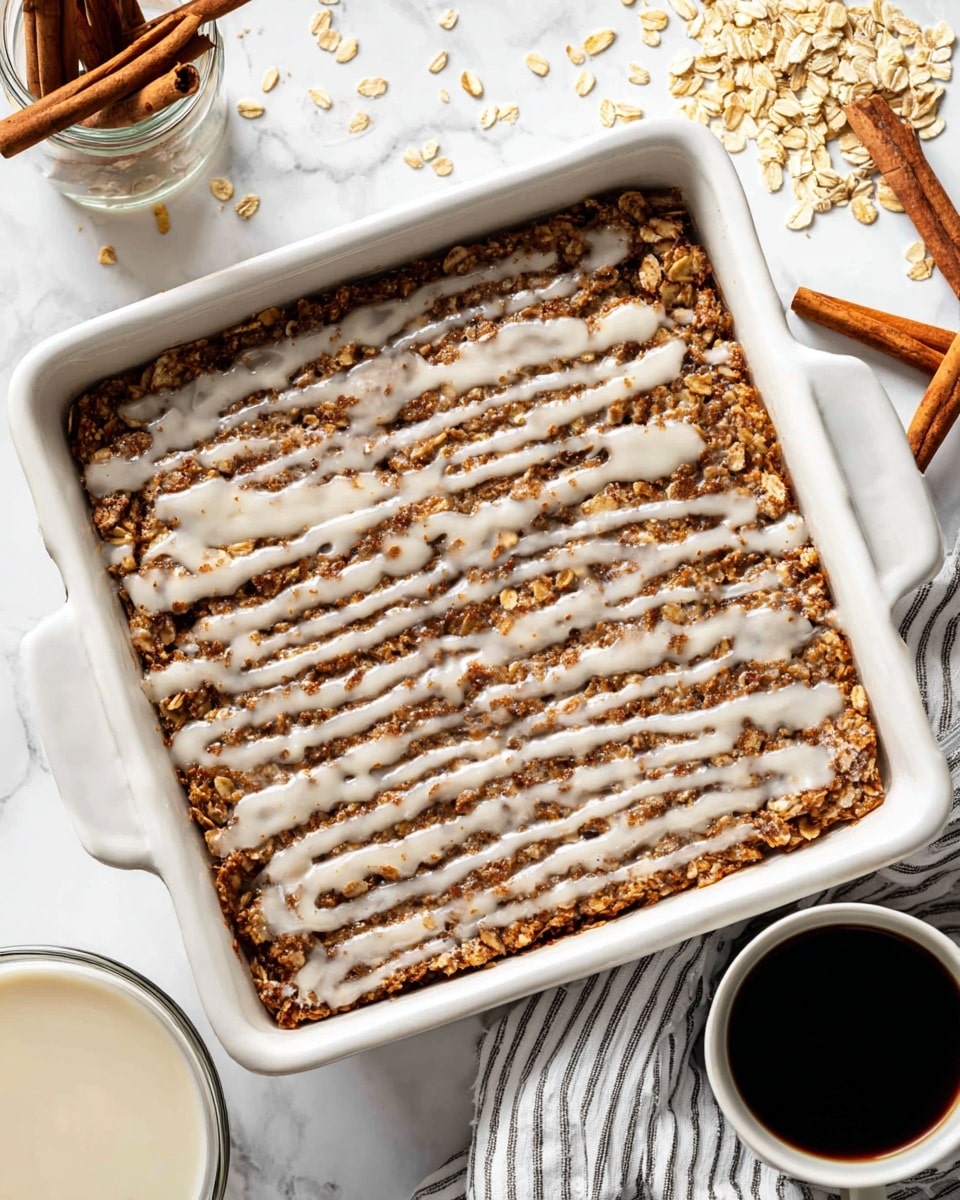 A white square baking dish filled with a baked oatmeal, topped with visible oats and a light drizzle of creamy white icing spread thinly in parallel lines across the surface, creating a mix of rough textured brown and beige oat clusters with smooth, shiny icing on top. The dish is placed on a white marbled surface with scattered oats around it, a glass jar holding several cinnamon sticks on the left, a small glass bowl with additional icing in the bottom left corner, a white cup filled with dark black coffee at the top right, and a white cloth with black stripes partially under the dish. photo taken with an iphone --ar 4:5 --v 7