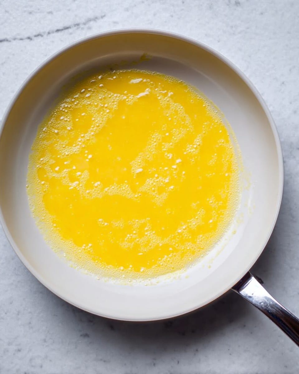 A close-up view of a white frying pan on a white marbled surface with a thin layer of bright yellow egg mixture cooking inside. The egg layer covers most of the pan bottom, slightly uneven with smooth and slightly bubbled texture, edges are soft and irregular. The pan handle is silver metal, extending to the right side of the image. Photo taken with an iphone --ar 4:5 --v 7