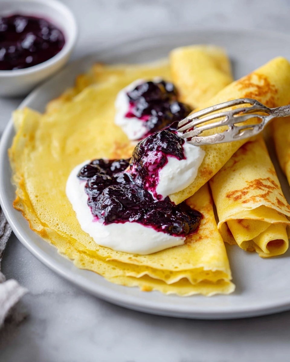 The image shows three thin yellow crepes folded into quarters, placed on a white plate. On top of the crepes, there is a dollop of white cream with a thick dark purple berry sauce spread over it. A fork holds a small rolled piece of crepe soaked with the same berry sauce and cream, resting on the plate. The background is a white marbled surface with a small bowl of extra dark purple berry sauce blurred in the foreground. Photo taken with an iphone --ar 4:5 --v 7