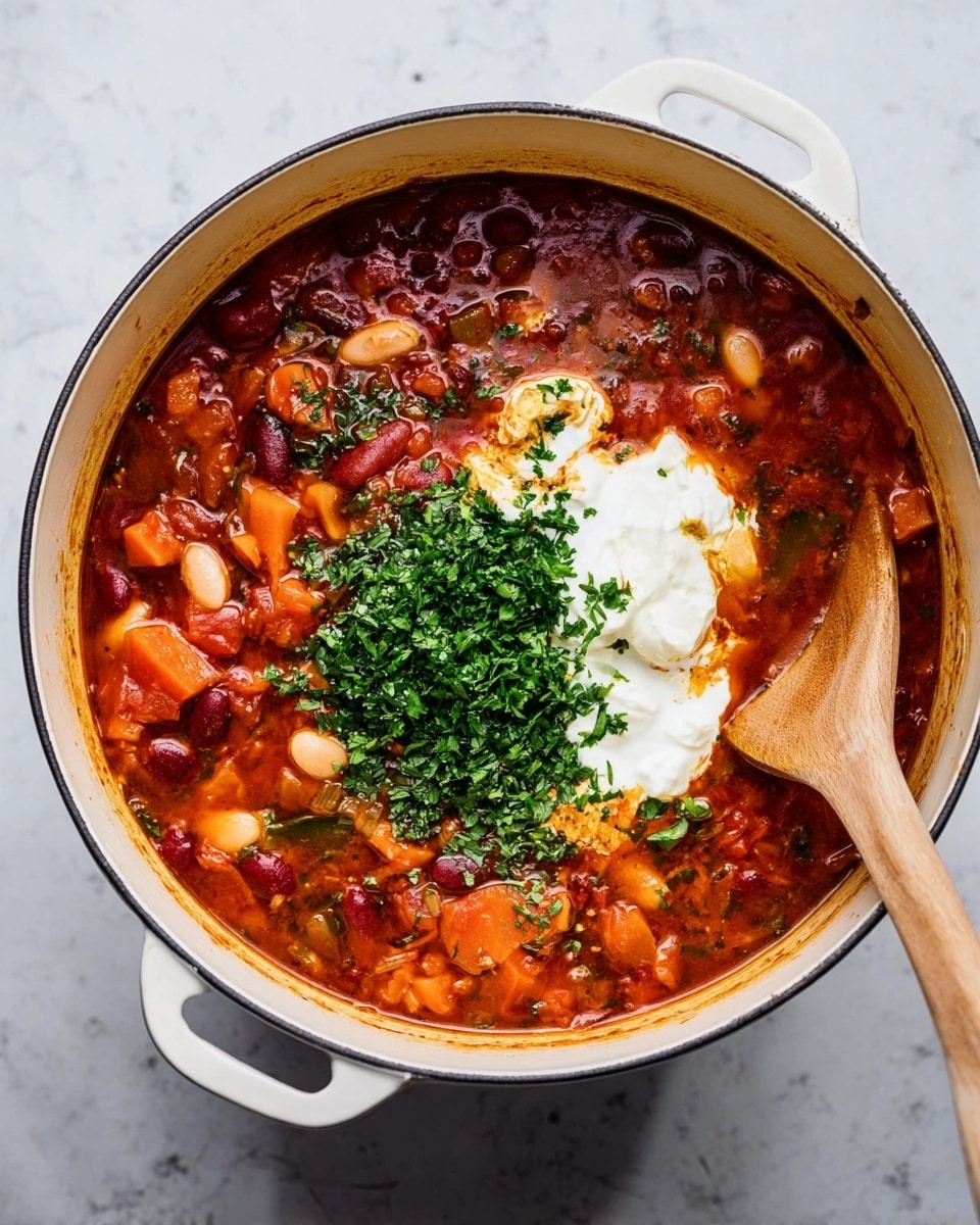 A white pot filled with chunky red soup showing visible pieces of orange and red vegetables and white beans in a rich broth. In the center of the soup, there is a dollop of smooth white cream next to a pile of chopped fresh green herbs. A wooden spoon is placed inside the pot resting against the edge. The pot sits on a white marbled surface. Photo taken with an iphone --ar 4:5 --v 7