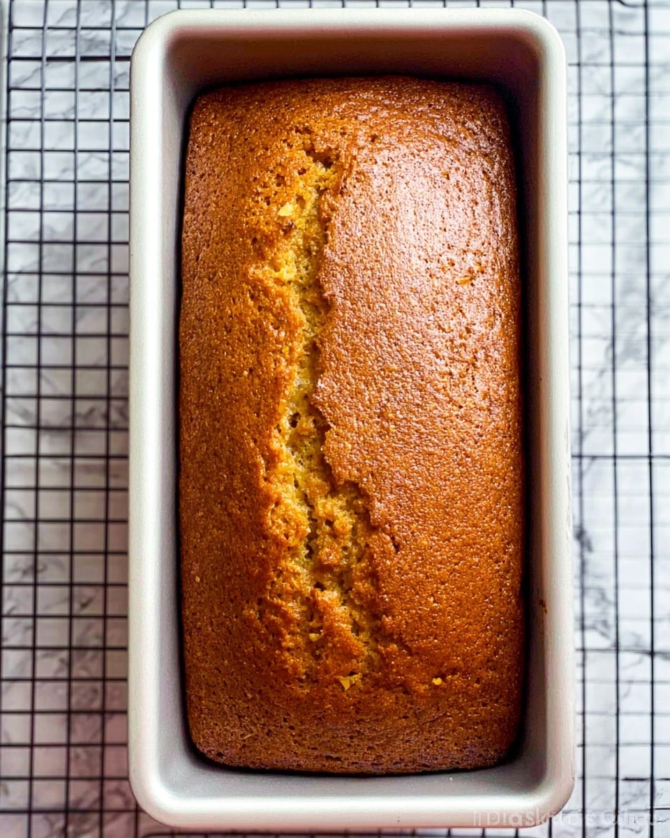 A single loaf of golden brown bread with a slightly cracked top surface sits in a rectangular white metal baking pan. The bread's texture looks soft and moist with small visible bits throughout. The pan is placed on a wire cooling rack, and the background shows a white marbled surface. Photo taken with an iphone --ar 4:5 --v 7