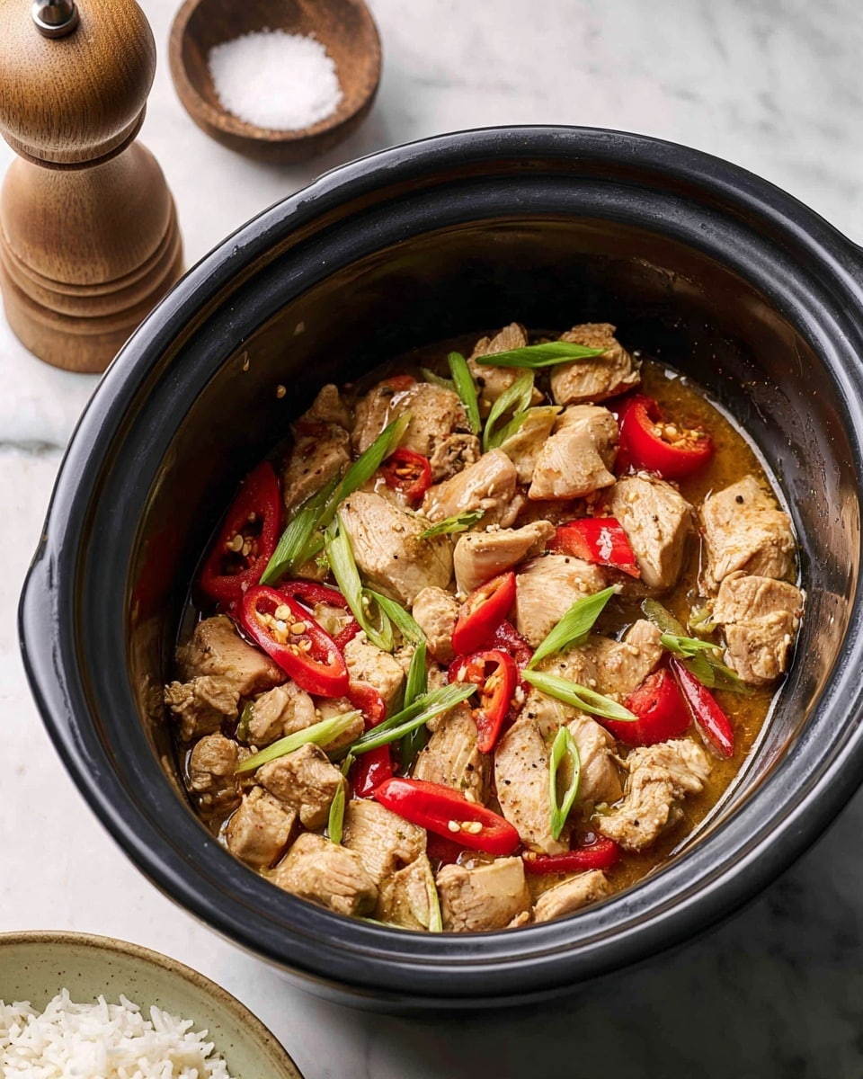 The image shows a black slow cooker filled with a cooked chicken dish. The dish has several chunky light brown chicken pieces cooked in a light brown sauce. Mixed in are sliced red chili peppers, red bell pepper pieces, and green onions cut into long sections. The slow cooker is placed on a white marbled surface, with a wooden pepper grinder and a bowl of salt nearby. On the lower side, part of a white plate with cooked white rice is visible. Photo taken with an iphone --ar 4:5 --v 7