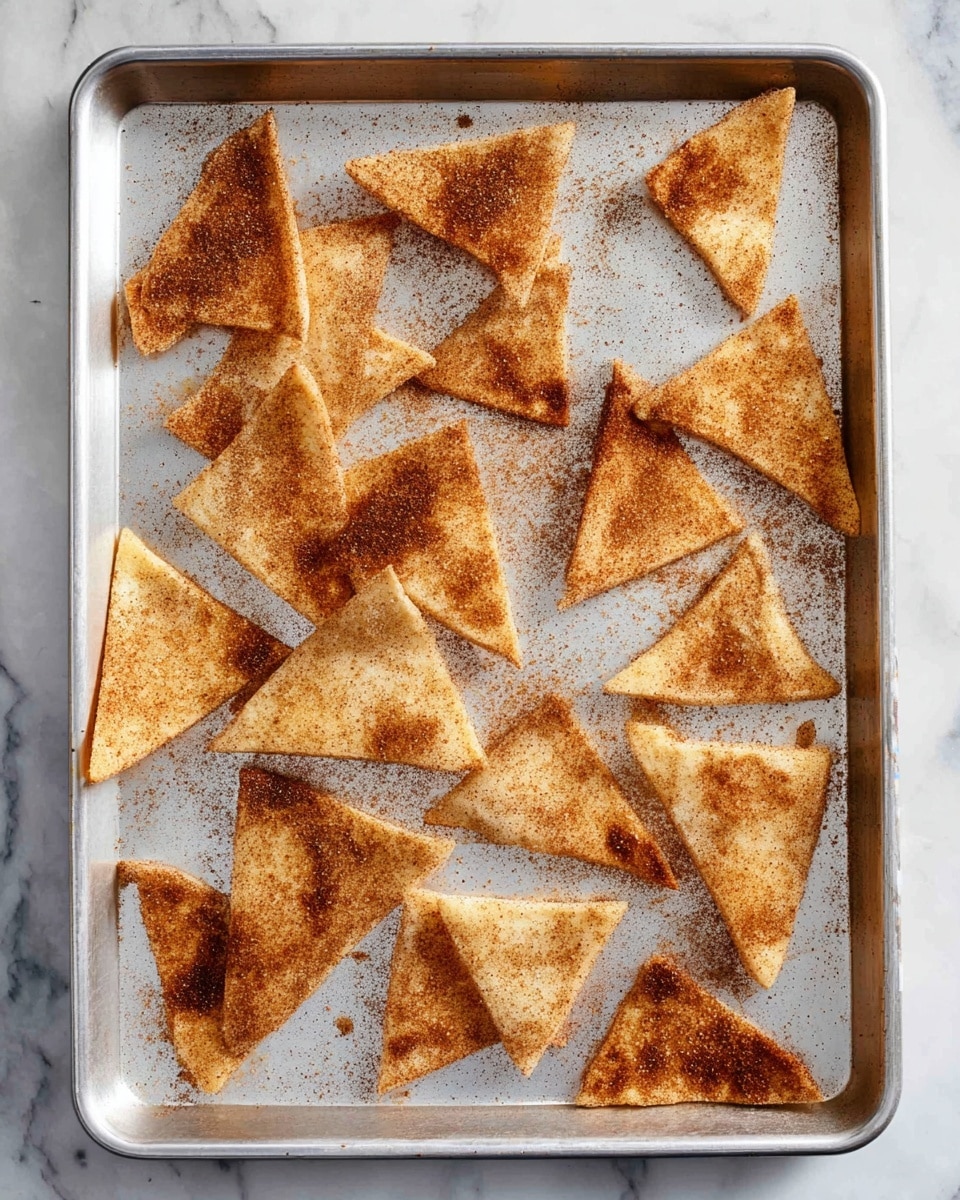 A silver baking tray holds 17 golden-brown triangular chips, each dusted with a light layer of cinnamon and sugar, showing a crispy texture on their surface. The chips are scattered unevenly across the tray, some overlapping slightly, with a few small loose crumbs and patches of cinnamon powder around them. The tray rests on a white marbled surface, which adds a clean, bright background to the warm tones of the chips. photo taken with an iphone --ar 4:5 --v 7