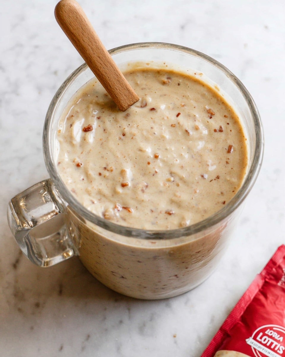 A clear glass cup filled with a creamy beige mixture that looks thick and textured with small brown bits evenly spread throughout. The cup has a clear handle holding a wooden spoon on its side. The surface where the cup sits shows a white marbled texture. Part of a red and white package is visible near the bottom right corner. Photo taken with an iphone --ar 4:5 --v 7