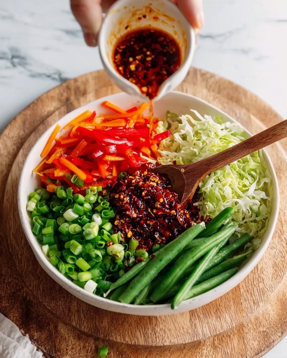 A white bowl sits on a wooden round board over a white marbled surface, filled with five fresh layers arranged in sections: chopped green onions on the bottom left, bright orange and red sliced bell peppers on the top left, finely shredded cabbage or lettuce on the top right, fresh green beans on the bottom right, and a dark, spicy chili mix in the center with seeds visible. A wooden spoon rests on the chili mix, and a woman's hand is pouring a dark red sauce with chili flakes from a small white cup over the bowl. The colors are vibrant and fresh, showing a mix of crisp vegetables and bold seasoning photo taken with an iphone --ar 4:5 --v 7