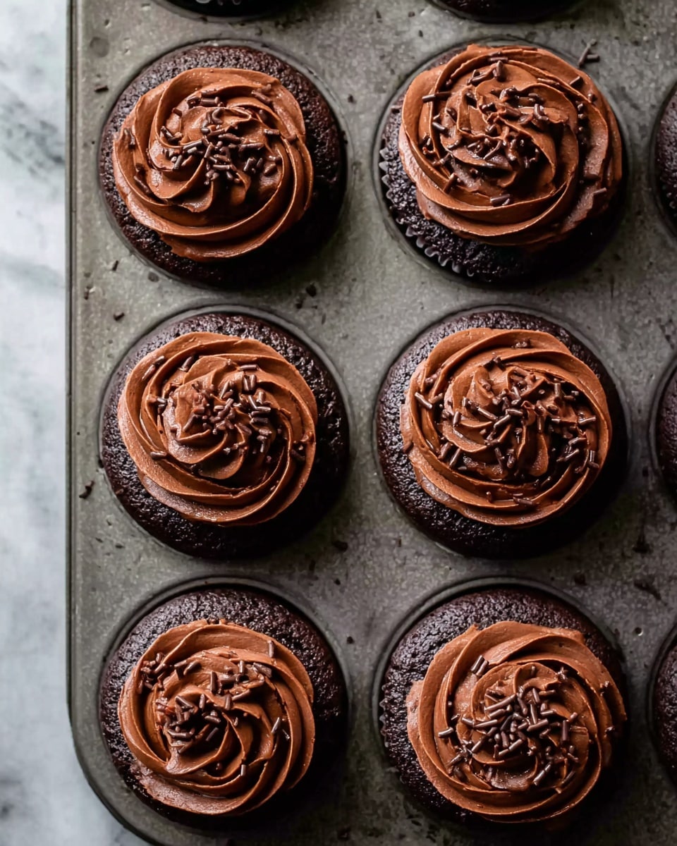 The image shows six chocolate cupcakes arranged in a metal cupcake tray. Each cupcake has two main layers: the base is a dark brown chocolate cake, and the top is a thick swirl of rich, shiny chocolate frosting, piped in a circular, floral pattern. The frosting is sprinkled with small chocolate shavings and tiny chocolate bits, adding texture and contrast to the smooth, creamy surface. The background surface is a white marbled texture. Photo taken with an iphone --ar 4:5 --v 7