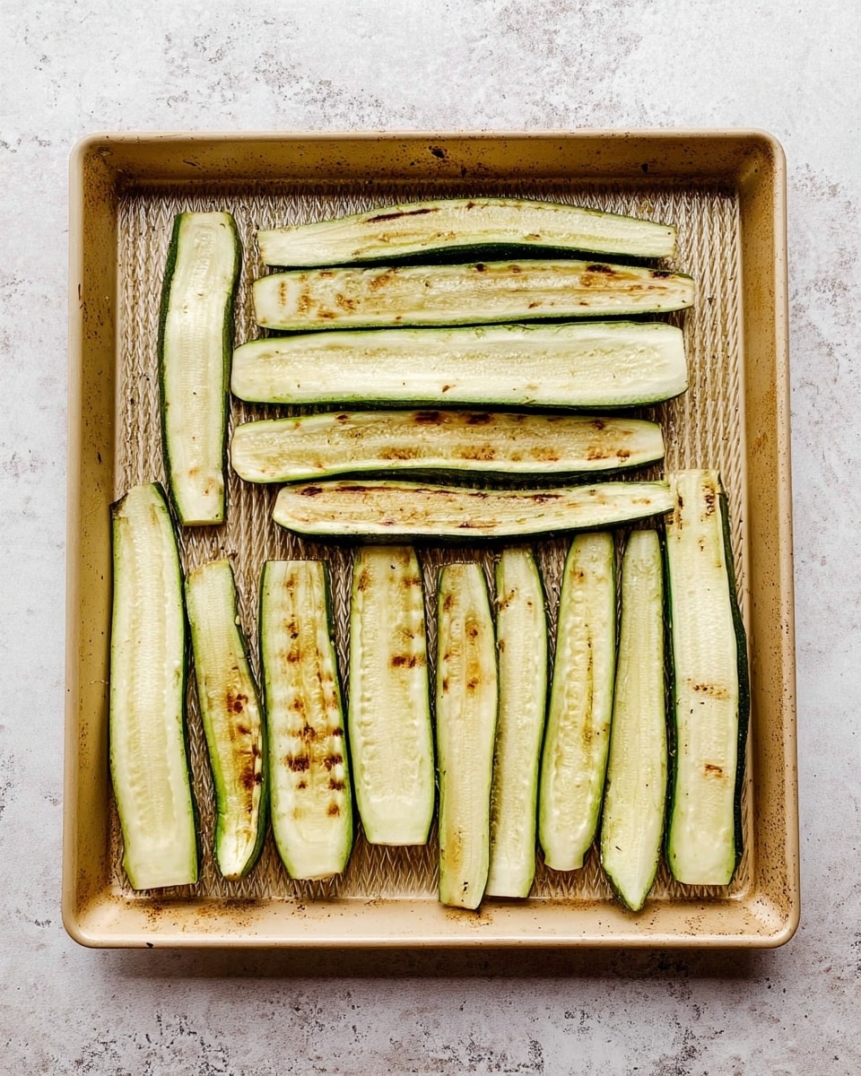 A baking tray filled with 11 long, thin zucchini slices arranged neatly in two rows on a white marbled surface. The zucchini slices are light green with grill marks of brown and golden patches visible on the surface. The tray itself has a textured pattern and is light tan in color, showing some slight signs of use. photo taken with an iphone --ar 4:5 --v 7